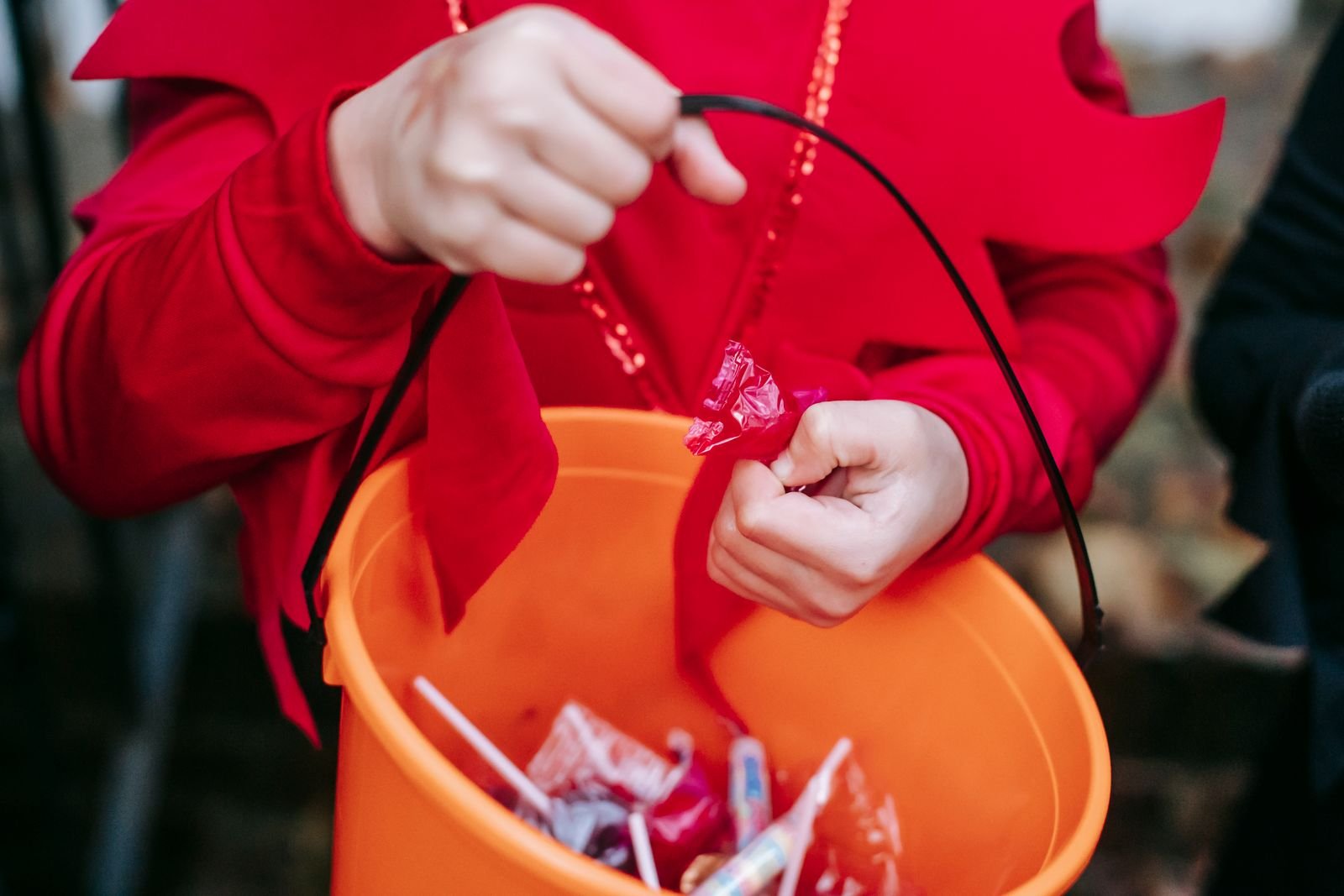 Child in a red costume holding an orange trick-or-treat bucket filled with wrapped candy